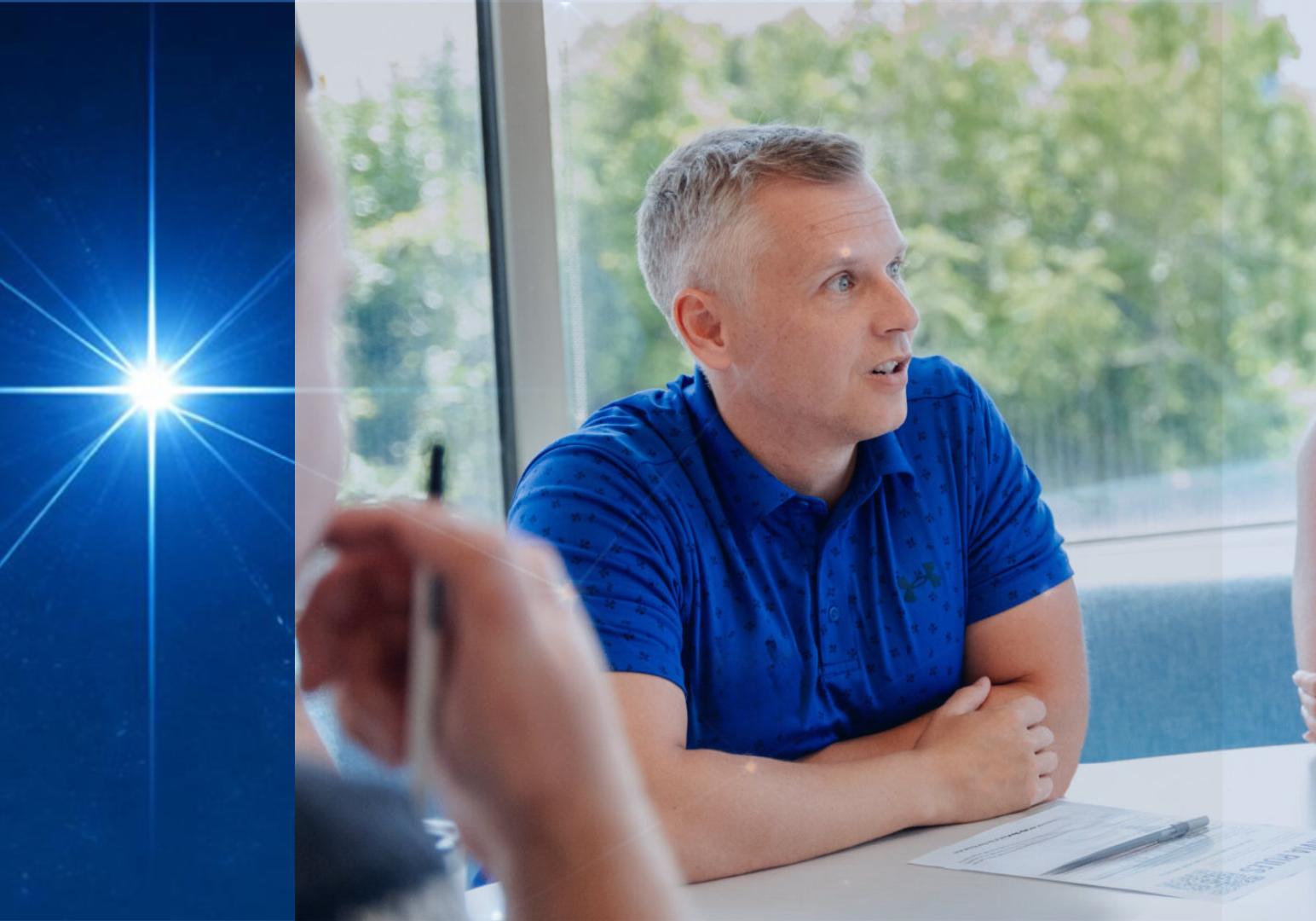 About Us Page Image - Carl and Lucida brightest star A man in a blue polo shirt speaks during a meeting at a conference table, beside a blue graphic panel with a bright starburst of light.